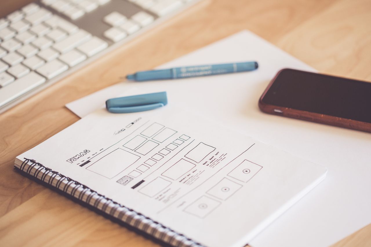 services-03 A neat workspace featuring a sketchpad with wireframes, a smartphone, and a keyboard on a wooden desk.